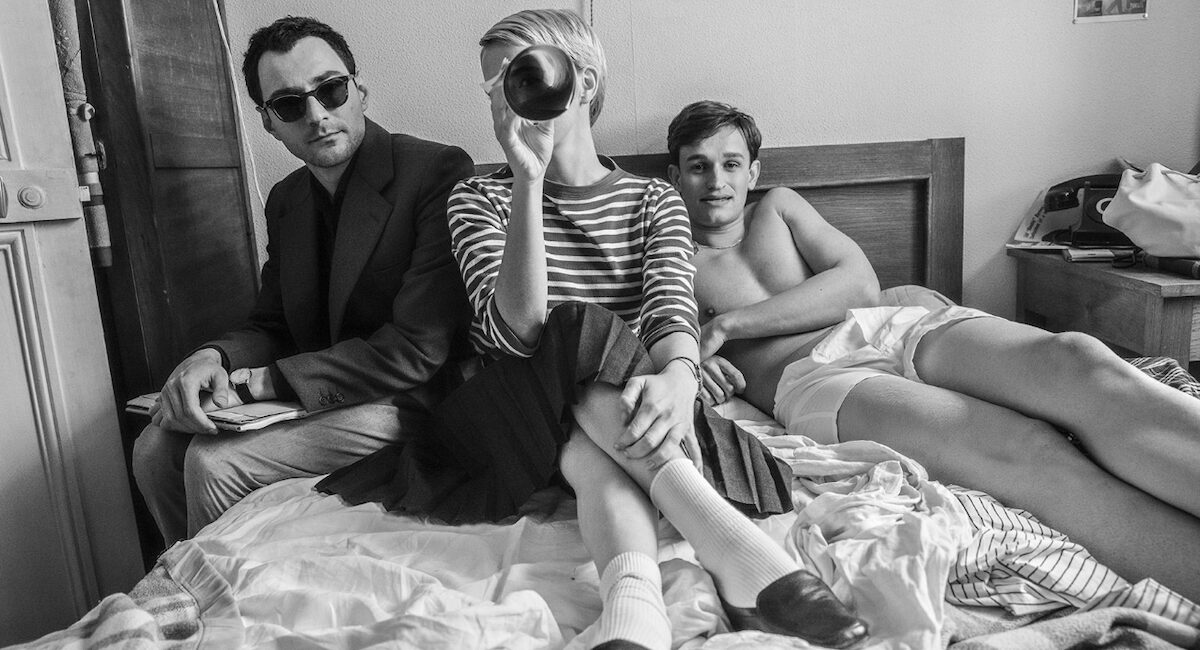 Woman in striped shirt and two men on bed, one shirtless